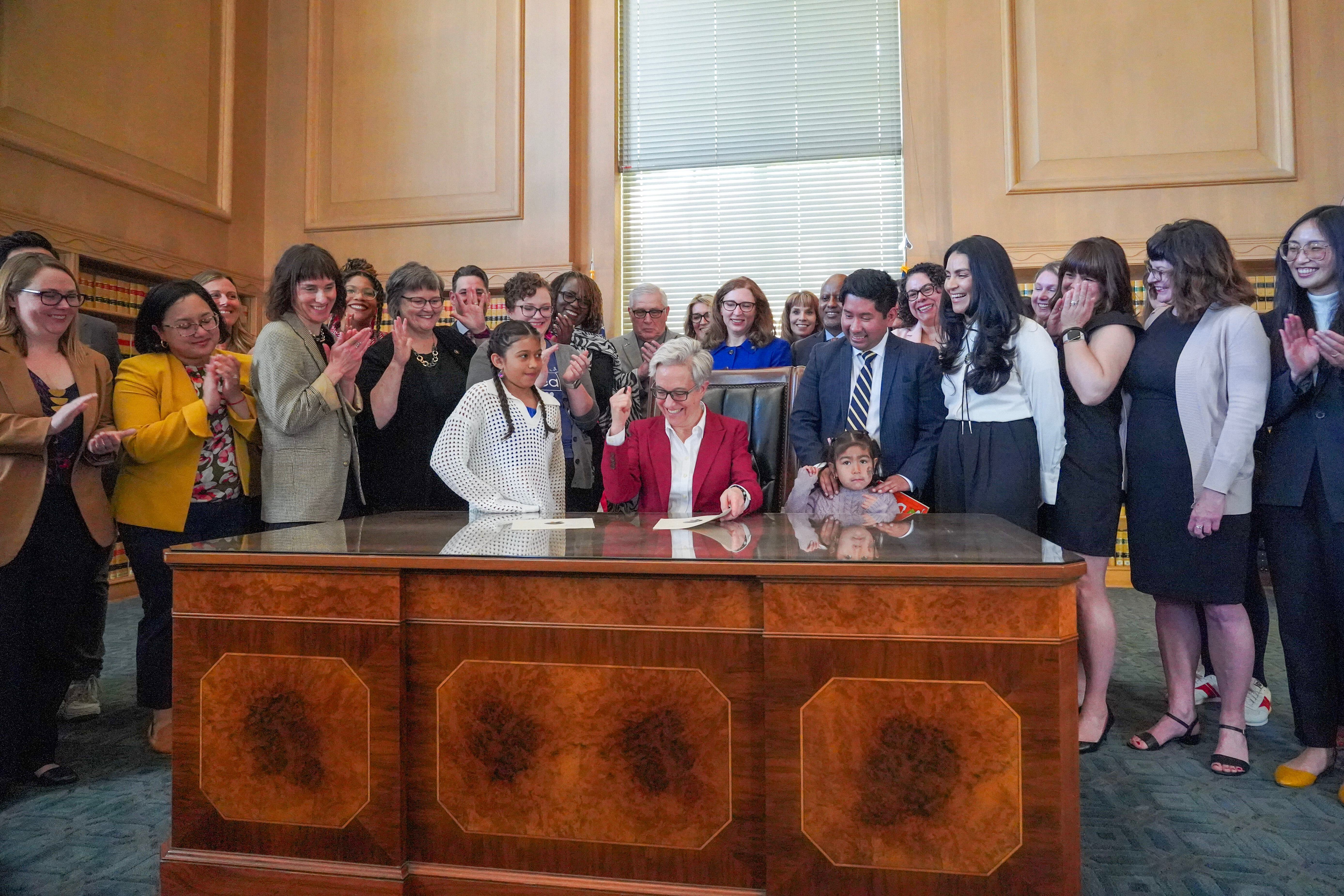 Gov. Tina Kotek is sitting at a large desk and celebrating after signing two summer learning bills, surrounded by state legislators and supporters.